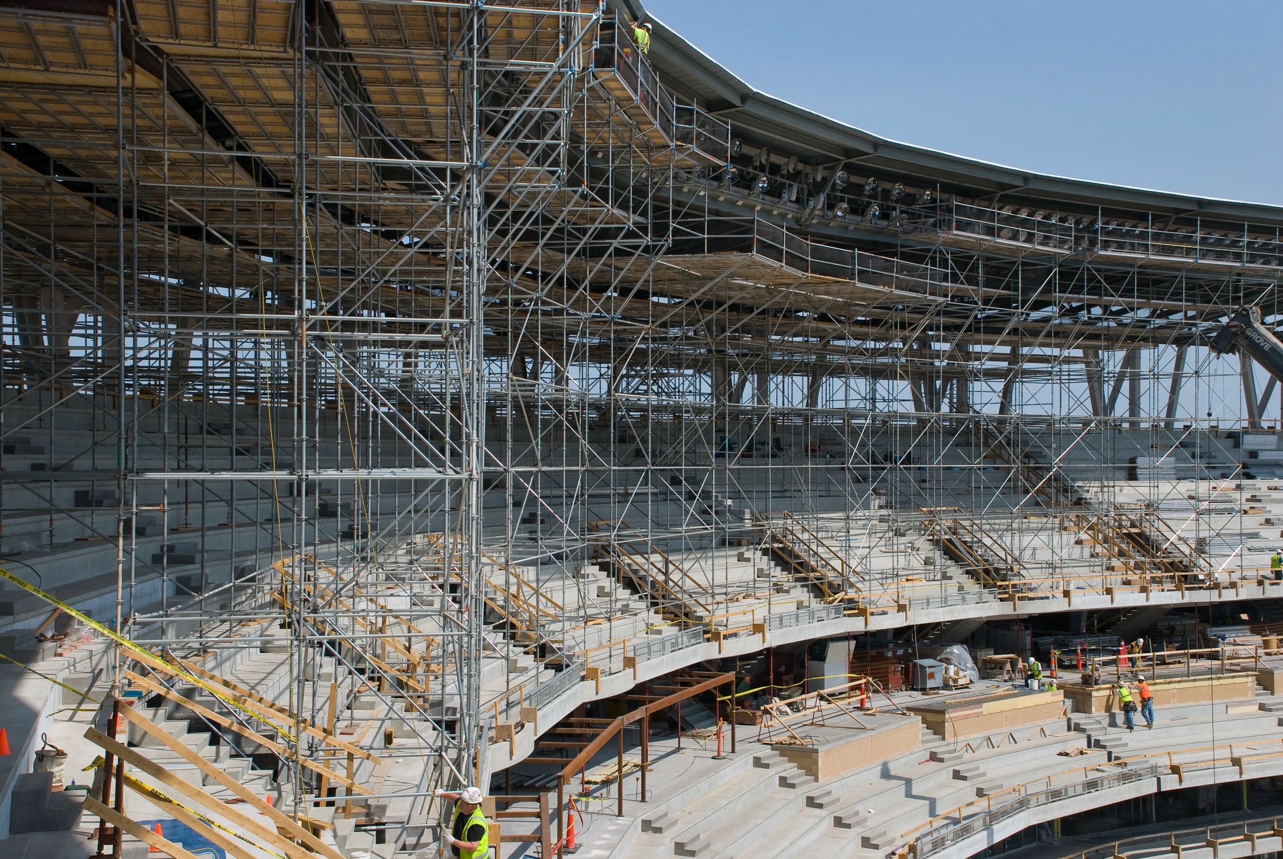 Construction workers build scaffolding inside a stadium under a clear sky.